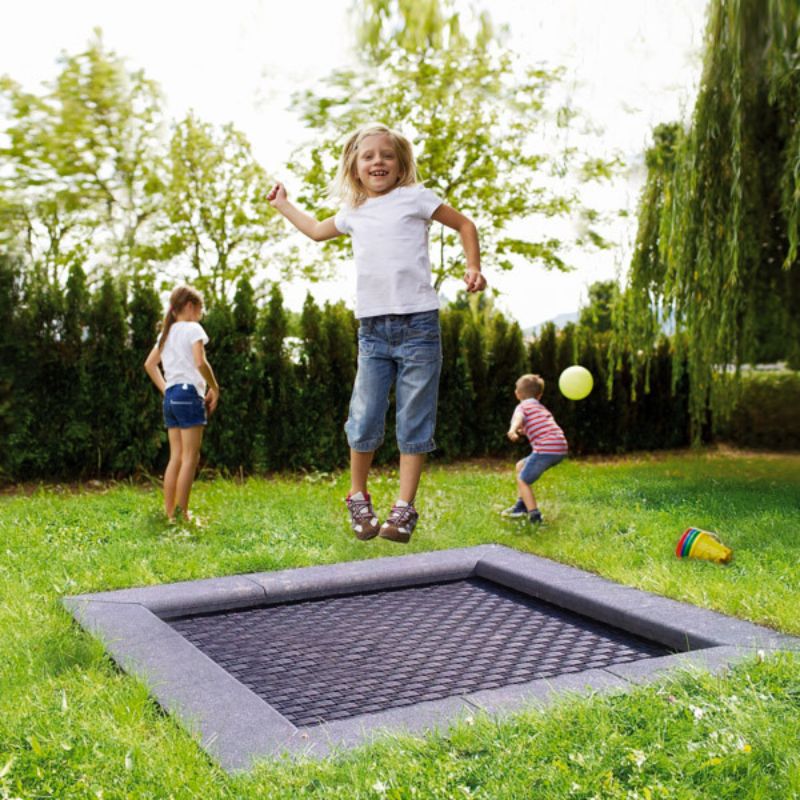 In eine Rasenfläche eingelassenes Bodentrampolin mit grauer Umrandung, darüber springt ein Mädchen, im Hintergrund spielen zwei Kinder